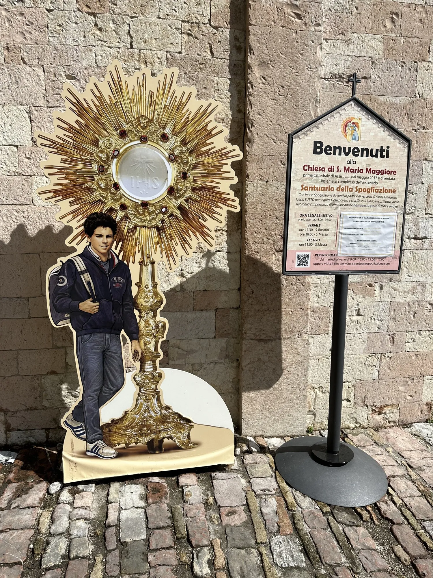 La sagoma di Carlo Acutis con un ostensorio dorato, accanto al cartello di benvenuto del Santuario della Spogliazione